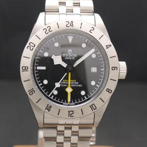 Puede incluir: Reloj de pulsera plateado con esfera negra, marcadores de hora y agujas blancas. El reloj tiene una correa de acero inoxidable y un bisel con números. La esfera muestra la marca "TUDOR GENÈVE" y "200m 660ft CHRONOMETER OFFICIALLY CERTIFIED".