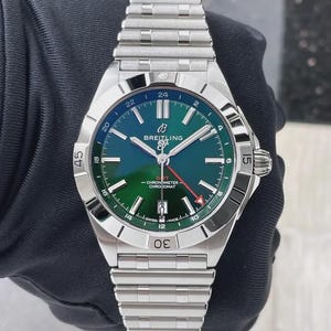 Op de afbeelding: Een Breitling Chronomat horloge van roestvrij staal met een groen en blauw wijzerplaat. Het horloge heeft een zilveren lunette met zwarte markeringen en een zilveren band. De wijzerplaat toont het Breitling-logo en de tekst "GMT Chronometer Chronomat".