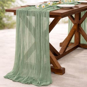 May include: A rustic wooden table is set for a gathering, adorned with a sage green table runner. White plates and daisy accents complement the decor. The table's legs are a dark brown, creating a warm, inviting atmosphere.