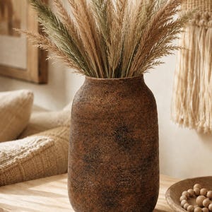 May include: A cylindrical vase with a dark brown, textured, weathered finish containing a large arrangement of tall, feathery, beige and light green pampas grass stems. The vase has a wide, rounded body and a slightly narrower opening at the top.