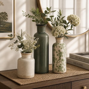 May include: Three decorative vases of varying sizes and styles, each holding greenery and flowers. The vases are placed on a woven mat atop a wooden surface. One vase is a solid, muted green, another is a cream color, and the third is patterned with green floral designs.