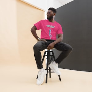 May include: A bright pink short-sleeved t-shirt featuring the word FOCUS printed in a stylized font above a graphic illustration of a person in a yoga pose. The shirt is paired with dark gray jeans and white athletic sneakers.