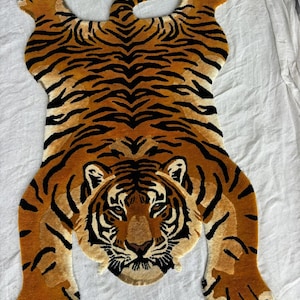 May include: A tufted rug shaped like a tiger, featuring a vibrant orange and brown coat with distinct black stripes. The design depicts the tiger in a sprawling position, showcasing a detailed face with white accents around the ears and paws.