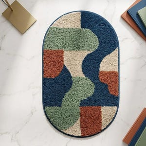 Puede incluir: Una alfombra de forma ovalada con una superficie texturizada y copetuda que presenta un patrón abstracto de formas entrelazadas en azul marino profundo, verde salvia, naranja terracota y crema. La alfombra tiene un borde azul marino sólido.