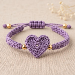 May include: A purple crochet heart bracelet featuring a central heart-shaped motif made of yarn. The band is constructed from braided purple cord with four small, polished gold-tone metal beads integrated into the design and an adjustable sliding knot.