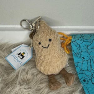 Könnte beinhalten: Ein beigefarbener Plüsch-Erdnuss-Schlüsselanhänger mit lächelndem Gesicht, braunen Kordbeinen und einem silbernen Metallverschluss. Ein weißes Jellycat Amuseables-Etikett ist befestigt. Daneben befindet sich ein Teil einer blauen Stofftasche mit schwarzem Muster.