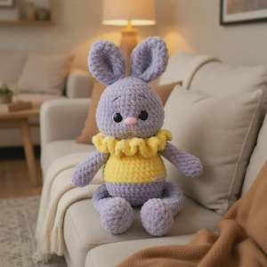 May include: A handmade crochet bunny doll in shades of lavender, yellow, and pink. The bunny has large ears, a yellow ruffled collar, and a two-tone body. The doll is sitting on a light-colored couch, suggesting a cozy, home-like setting.