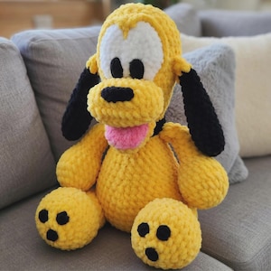 Könnte beinhalten: Ein handgefertigtes Plüschtier von Pluto, dem Disney-Zeichentrickhund. Das Spielzeug ist hauptsächlich gelb mit weißen Akzenten um die Augen, schwarzen Ohren und einer rosa Zunge. Das Plüschtier sitzt auf einem grauen Sofa.