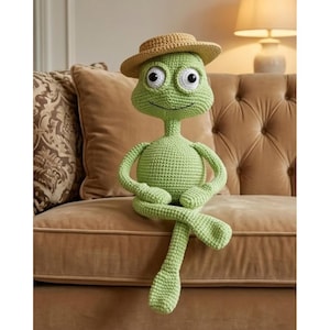 May include: A handmade, light green crocheted alien doll with large, white-eyed features and a small, tan straw hat. The alien is seated on a brown sofa, its arms and legs crossed. A whimsical, soft toy.