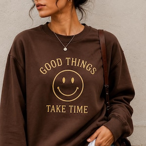 May include: A dark brown crewneck sweatshirt featuring a gold graphic of a smiley face centred between the text GOOD THINGS and TAKE TIME. The garment has long sleeves with ribbed cuffs and a ribbed hem, worn over a white shirt with blue jeans.