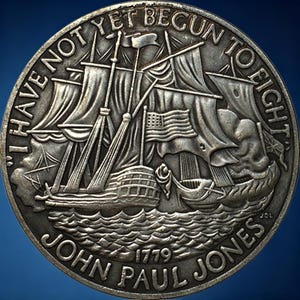 May include: A silver-colored coin featuring a detailed depiction of a sailing ship with billowing sails and the American flag. The coin is inscribed with the text "I HAVE NOT YET BEGUN TO FIGHT" and "JOHN PAUL JONES 1779".