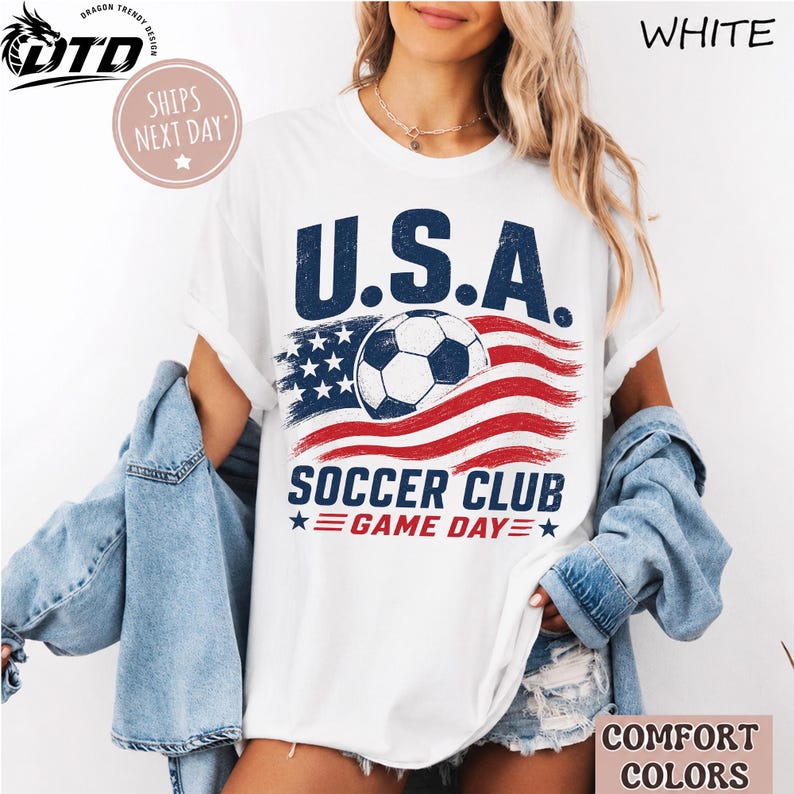 K&ouml;nnte beinhalten: Ein wei&szlig;es Kurzarm-T-Shirt mit einem Grafikdruck und dem Text U.S.A. SOCCER CLUB GAME DAY. Das Design zeigt einen Fu&szlig;ball, der &uuml;ber einem stilisierten amerikanischen Flaggenmotiv in Rot, Wei&szlig; und Blau mit einer abgenutzten Textur zentriert ist.