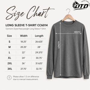 K&ouml;nnte beinhalten: Ein anthrazitfarbenes Langarm-T-Shirt auf einem Holzkleiderb&uuml;gel neben einer Gr&ouml;&szlig;entabelle mit der &Uuml;berschrift Size Chart, die Breiten und L&auml;ngen in Zoll f&uuml;r die Gr&ouml;&szlig;en S bis 3XL des kleidungsst&uuml;ckgef&auml;rbten Langarm-T-Shirts CC6014 auflistet.