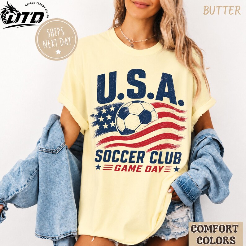 K&ouml;nnte beinhalten: Ein buttergelbes Kurzarm-T-Shirt mit einem Grafikdruck und dem Text U.S.A. SOCCER CLUB GAME DAY. Das Design zeigt einen Fu&szlig;ball, der &uuml;ber einem abgenutzten amerikanischen Flaggensymbol in den Farben Rot, Wei&szlig; und Blau zentriert ist.