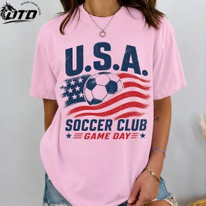 K&ouml;nnte beinhalten: Ein hellrosa T-Shirt mit kurzen &Auml;rmeln und einer verwaschenen Grafik eines Fu&szlig;balls, der &uuml;ber einer stilisierten amerikanischen Flagge zentriert ist. Der Text lautet U.S.A. SOCCER CLUB GAME DAY in dunkelblauer und roter Schrift.