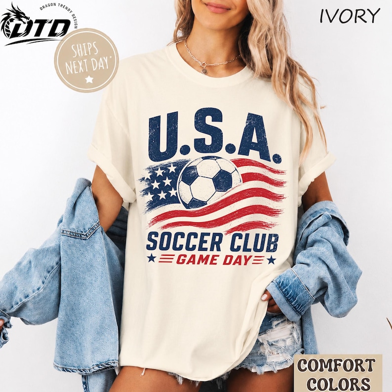 K&ouml;nnte beinhalten: Ein elfenbeinfarbenes Kurzarm-T-Shirt mit einem Grafikdruck und dem Text U.S.A. SOCCER CLUB GAME DAY. Das Design zeigt einen Fu&szlig;ball, der &uuml;ber einer stilisierten amerikanischen Flagge mit abgenutzter Textur in den Farben Rot, Wei&szlig; und Blau liegt.