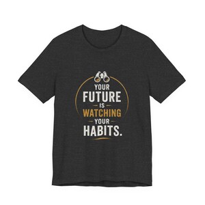 以下が含まれることがあります： 「YOUR FUTURE IS WATCHING YOUR HABITS」の文字が入った濃いグレーのTシャツ。デザインには双眼鏡が付いた金色の円が含まれています。テキストは白と金です。