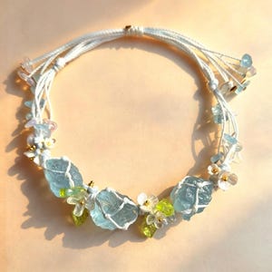 May include: A delicate necklace featuring large, light blue aquamarine stones, accented with small white flowers, green leaves, and gold beads. The stones are held in place with white macrame cord, creating a bohemian style.