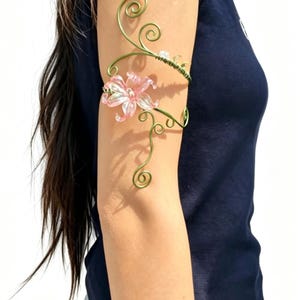 May include: A delicate, gold-toned arm cuff featuring a pink flower and swirling vine design. The floral accent is a translucent pink with white detailing. The cuff wraps around the upper arm, creating an elegant, nature-inspired accessory.