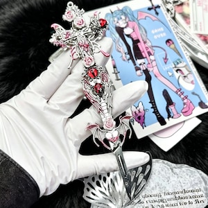 Könnte beinhalten: Ein silberfarbener Brieföffner in Kreuzform mit roten und rosa Akzenten. Der Öffner hat Rosen- und Flügeldetails sowie ein herzförmiges Design. Im Hintergrund ist eine farbenfrohe Illustration mit den Worten "GAME OVER" zu sehen.