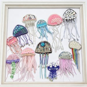 May include: A collection of handcrafted jellyfish brooches in a variety of colors and designs. Each brooch features intricate beadwork, sequins, and delicate fringe, displayed in a white framed presentation.