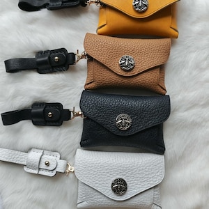 May include: Four small rectangular pouches in mustard yellow, tan, black, and white, each featuring a textured surface, a flap closure secured with a silver-toned bee emblem, and a matching leather strap attached by a gold-toned metal swivel clasp.