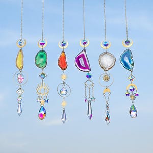 May include: A collection of hanging suncatchers with colorful agate slices, crystals, and gold-toned metal accents. Each suncatcher features a unique agate slice in shades of yellow, green, red, purple, white, and blue, suspended from a chain.
