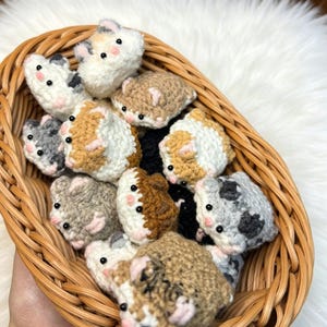 以下が含まれることがあります： 手作りのかぎ針編みモルモットのおもちゃがたくさん入った柳のバスケット。モルモットは白、茶色、グレー、黒など様々な色があり、ピンクの耳と黒いビーズの目があります。バスケットはふわふわの白い表面に置かれています。