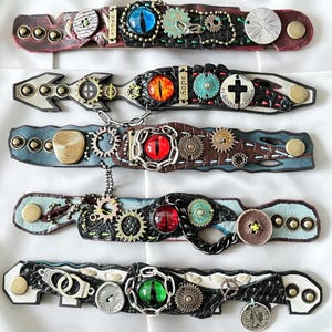 May include: Five handcrafted leather bracelets with steampunk and gothic elements. Each bracelet features unique designs with gears, eye-shaped cabochons, metal accents, and decorative stitching. Colors include brown, black, blue, and red.
