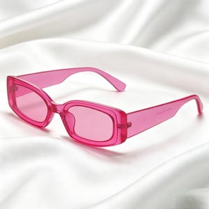 May include: A pair of bright pink sunglasses with a rectangular frame. The sunglasses have a translucent appearance and are set against a white, silky background. The text "IN CLOVE WITH" is printed on the arm of the glasses.
