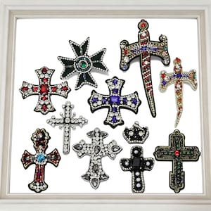 May include: A collection of decorative cross-shaped brooches with various designs. The brooches feature an array of colors, including red, green, blue, and clear crystals. Some crosses have a sword-like design, while others are adorned with pearls and black accents.