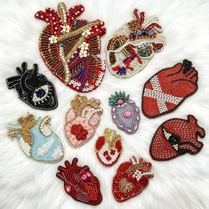 Puede incluir: Una colección de once parches con forma de corazón anatómico elaborados con intrincados abalorios, lentejuelas e hilos metálicos en varias paletas de colores, incluyendo rojo, oro, negro, azul claro y rosa pastel, dispuestos sobre una superficie blanca texturizada.