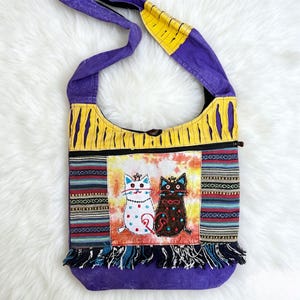 May include: A purple and yellow shoulder bag with a cat design. The bag features a colorful embroidered panel with two cat figures, a zipper closure, and a fringed bottom. The strap is purple and yellow.