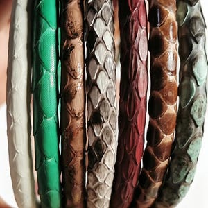 May include: A collection of seven leather bracelets in various colors, including white, green, brown, gray, red, and teal. The bracelets have a textured, scale-like pattern, and are held in a hand.