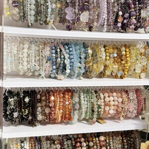 May include: A display of beaded bracelets in various colors, including purple, blue, yellow, and pink. The bracelets are arranged on white shelves, showcasing a variety of gemstone and crystal designs. The beads range in size and texture, creating a visually appealing collection.