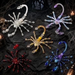 Puede incluir: Cinco figuras decorativas de escorpión hechas con cuentas y cristales de varios colores. La colección incluye un escorpión blanco, uno morado y azul, uno amarillo y dorado, uno rojo y negro, y uno azul y morado, cada uno con cola curva.