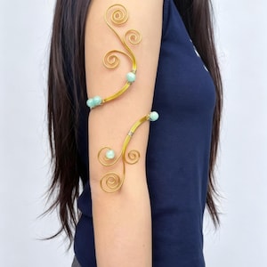 May include: Gold-toned arm cuff with a swirling vine design, adorned with small, light blue beads. The cuff wraps around the upper and lower arm, creating an elegant, nature-inspired accessory. The jewelry is shown on a person wearing a navy blue top.