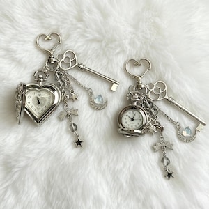 Peut inclure: Deux porte-clés argentés avec des montres de poche, des clés et des breloques célestes. L'un des porte-clés présente une montre en forme de cœur, tandis que l'autre a une montre ronde. Les deux ont des accents d'étoiles, de lune et de nœuds.