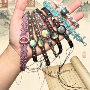 Peut inclure: Plusieurs bracelets en macramé faits à la main, de couleurs variées, notamment violet, marron et noir. Chaque bracelet est orné d'une pierre précieuse ou d'un élément décoratif central. Les bracelets sont présentés sur un fond de peinture d'inspiration asiatique.