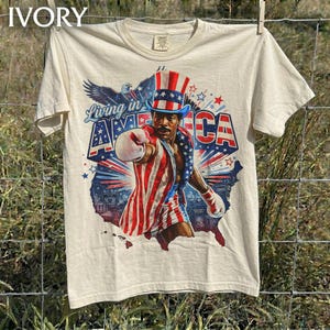 May include: A cream-colored short-sleeve t-shirt featuring a graphic of a person dressed as Uncle Sam in red, white, and blue attire. The design includes a bald eagle, a map of the United States, and the text Living in America in blue lettering.