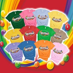 Könnte beinhalten: Eine Sammlung von elf kurzärmeligen T-Shirts in verschiedenen Unifarben, darunter Grün, Pink, Hellblau, Gelb, Rot, Braun, Orange und Lavendel. Jedes T-Shirt zeigt das Skittles-Logo, das in einem Oval auf der Brust aufgedruckt ist.