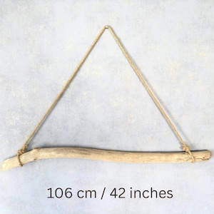 Op de afbeelding: Een natuurlijk drijfhouten plank, 106 cm lang, opgehangen aan een jute touw. Het lichtgekleurde hout heeft een gebogen vorm en is aan elk uiteinde geknoopt om het touw vast te zetten. De plank hangt tegen een lichtgrijze, getextureerde achtergrond.