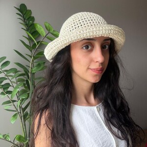 Puede incluir: Un sombrero de pescador de ganchillo color crema con un patrón de tejido abierto en la copa y un ala sólida y estrechamente tejida. El sombrero está hecho de hilo y presenta una parte superior redondeada con un ala ancha e inclinada.