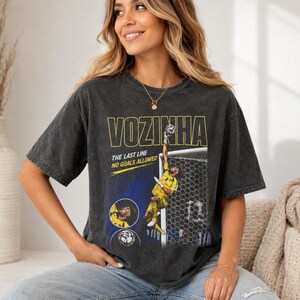 Peut inclure: Un t-shirt gris anthracite &agrave; manches courtes avec une impression graphique d'un gardien de but de football en maillot jaune. Le texte indique VOZINHA, THE LAST LINE et NO GOALS ALLOWED. Le design inclut un filet de but et une image circulaire.