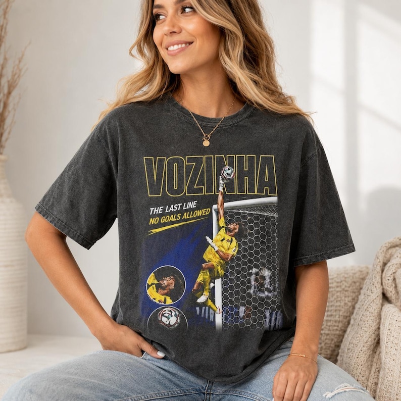 Peut inclure: Un t-shirt gris anthracite &agrave; manches courtes avec une impression graphique d'un gardien de but de football en maillot jaune. Le texte indique VOZINHA, THE LAST LINE et NO GOALS ALLOWED. Le design inclut un filet de but et une image circulaire.