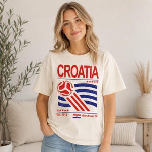 Puede incluir: Una camiseta de manga corta color crema con la palabra CROATIA en texto rojo sobre un gr&aacute;fico de un bal&oacute;n de f&uacute;tbol rojo y l&iacute;neas onduladas azules. Debajo del gr&aacute;fico, el texto dice Est. 1912, World Cup '26 e incluye una peque&ntilde;a bandera croata.