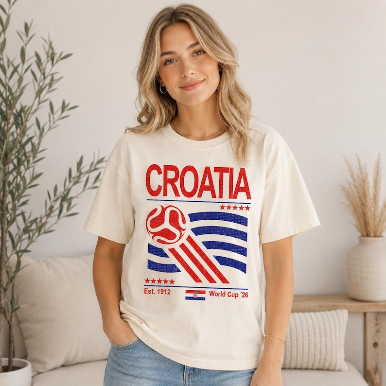 Puede incluir: Una camiseta de manga corta color crema con la palabra CROATIA en texto rojo sobre un gr&aacute;fico de un bal&oacute;n de f&uacute;tbol rojo y l&iacute;neas onduladas azules. Debajo del gr&aacute;fico, el texto dice Est. 1912, World Cup '26 e incluye una peque&ntilde;a bandera croata.