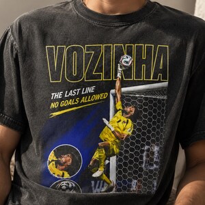 Peut inclure: Un t-shirt noir anthracite avec un graphisme repr&eacute;sentant un gardien de but en uniforme jaune sautant pour attraper un ballon. Le texte indique VOZINHA, THE LAST LINE et NO GOALS ALLOWED. Le design inclut un encart circulaire du visage.