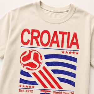 Puede incluir: Una camiseta de color crema con un estampado gr&aacute;fico en rojo y azul. El texto dice CROATIA en la parte superior, con un bal&oacute;n de f&uacute;tbol, l&iacute;neas onduladas, cinco estrellas, el texto Est. 1912, una peque&ntilde;a bandera croata y World Cup '26 abajo.