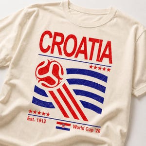 Puede incluir: Una camiseta de manga corta color crema con un estampado gr&aacute;fico desgastado. El dise&ntilde;o incluye la palabra CROATIA en rojo, un bal&oacute;n de f&uacute;tbol rojo estilizado, rayas horizontales azules, cinco estrellas rojas, el texto Est. 1912 y World Cup '26.
